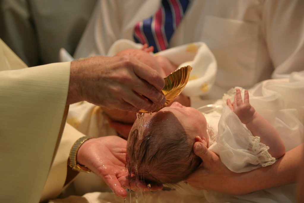Close up of baby girl being baptized. Hands of priest and God parents visible; water clearly visible flowing over baby's head. (Twin brother in background.)